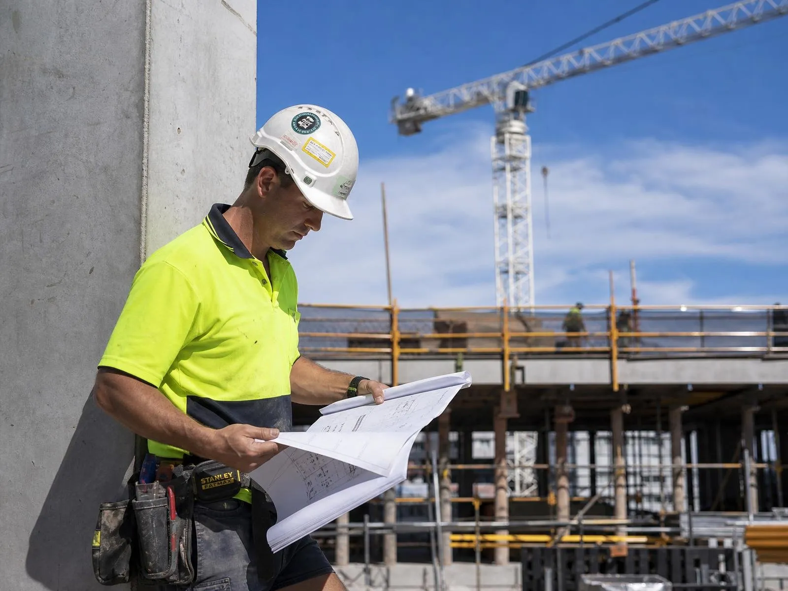 Australian mid-rise commercial construction supervisor reviewing plans against a concrete column